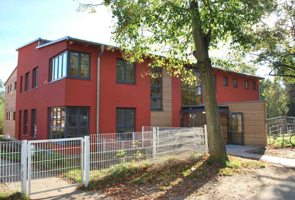 Ein zweistöckiges Haus mit roter Fassade, teilweise mit Holzelementen. Davor ein weißer Zaun und ein Baum.