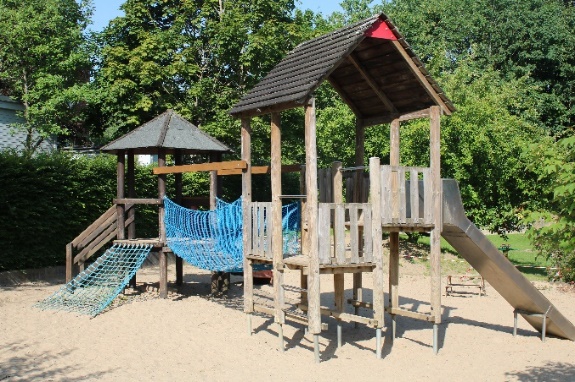 Spielplatz im Außenbereich mit Sandboden. Zwei offene Spielhäuser sind mit einer blauen Brücke verbunden. An einem Haus befindet sich eine Rutsche. Der Hintergrund ist begrünt.